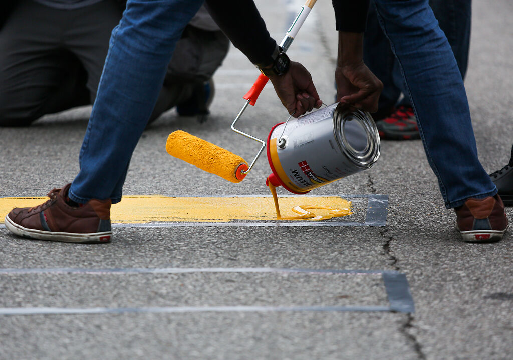 A protester pours paint while another spreads it with a roller in a taped outline that reads "Resign Hawley"